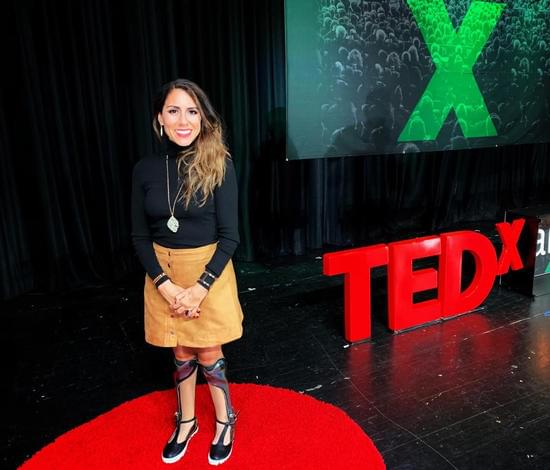 Photo of Estela Lugo, disability speaker standing on a stage ontop of the TEDx red dot carpet. Behind her is the TEDx Farmingdale logo. She is wearing a black turtleneck and camel brown , sued knee-length skirt. Estela is showing her bilateral leg braces.