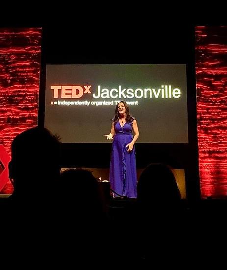 Photo of Lainie Ishbia, a disability speaker is standing on the TEDx stage wearing a long royal blue, sleevless dress. Behind her is a large screen with the TEDx Jacksonville logo displayed across it. On either side are two textured, uplit panels in red.