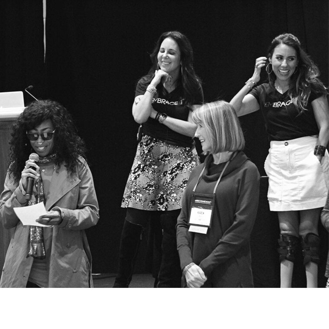 In this black and white photo, Lainie Ishbia and Estela Lugo are shown standing on stage during a conference. Infront of and below them are two female conference attendees. They are interacting during an excersise. The woman on the left is black woman with long curly black hair. She is wearing sunglasses a trenchcoat and is speaking into a micropone while reading from a sheet of paper in her other hnad. The woman on the right is a caucasian woman with blonde, shoulder-length hair. She is wearing a dark cowlneck sweater. Her head is turned to the woman next to hear as she smiles.