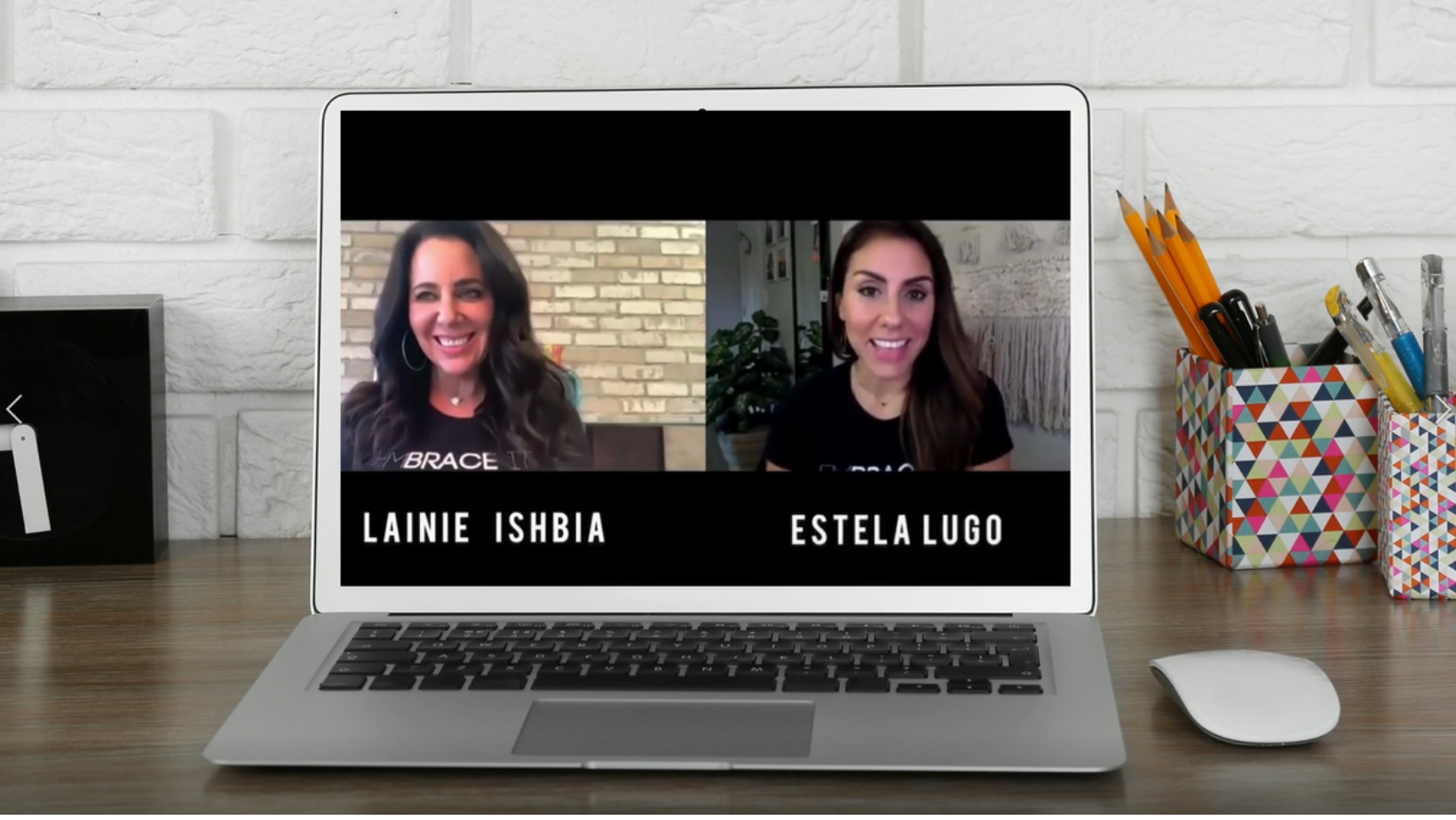 Photo of a silver laptop sitting on a dark wood desk with a virtual meeting screen showing Lainie Ishbia and Estela Lugo side-by-side with their names displayed below. begind the laptop is a white painted brick wall and colorful, geometric printed pencil boxes holding pencils and pens. A white computer mouse sits on the right side of the laptop.